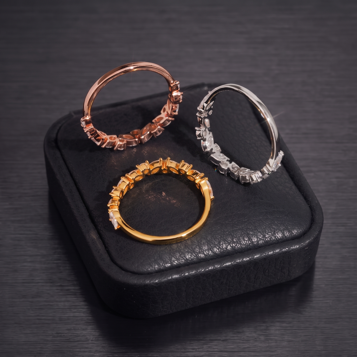 Rose gold, silver and gold marquise moissanite stackable eternity bands on black leather box over brushed black metal