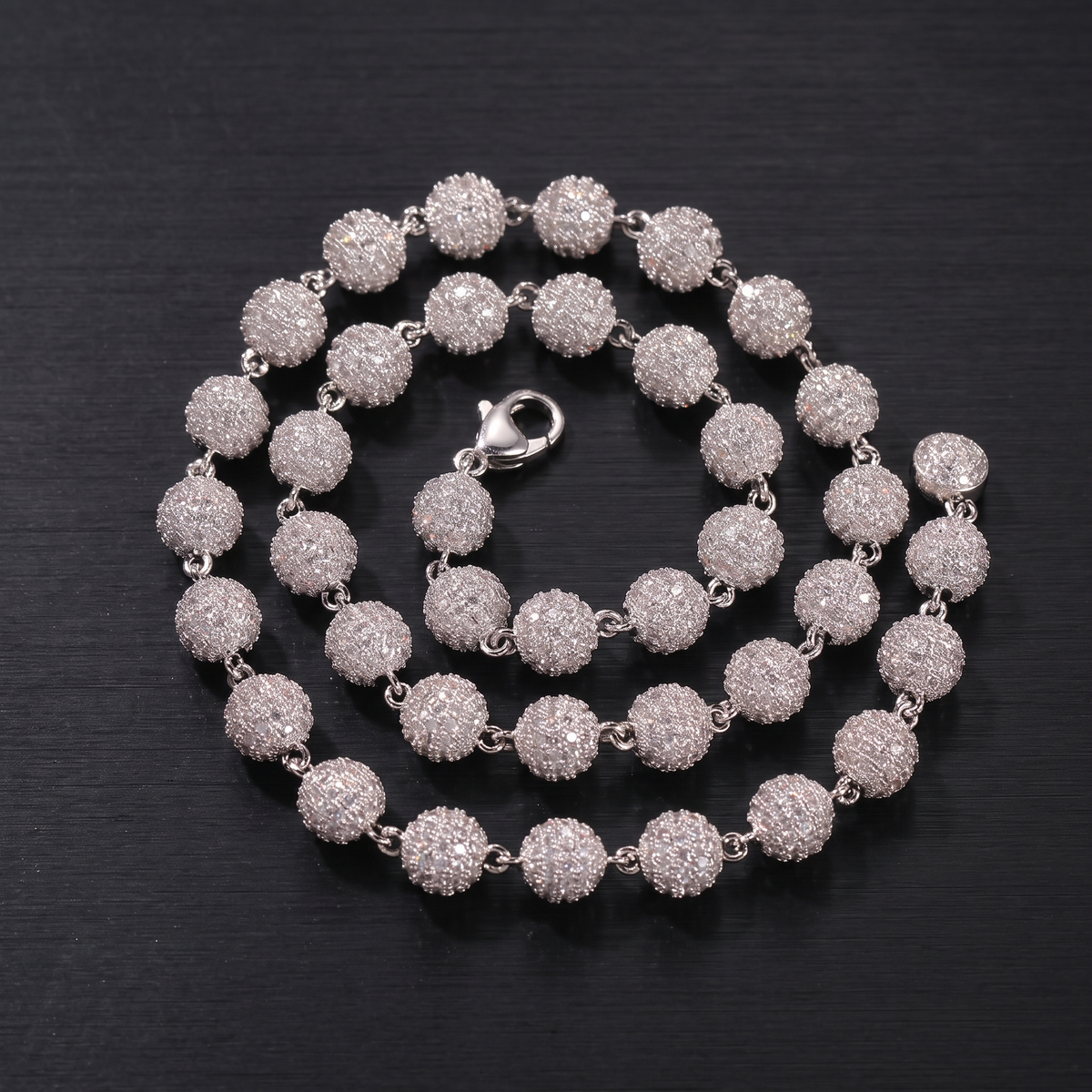 Silver 8mm iced out ball chain coiled on a black brushed steel surface
